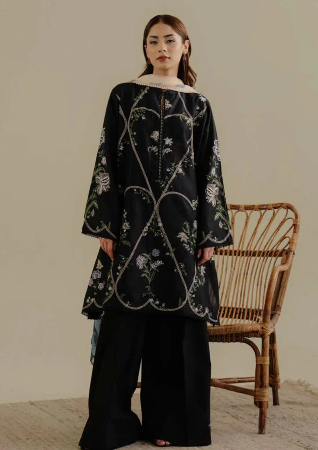 Woman wearing a black floral embroidered dress standing next to a wicker chair.