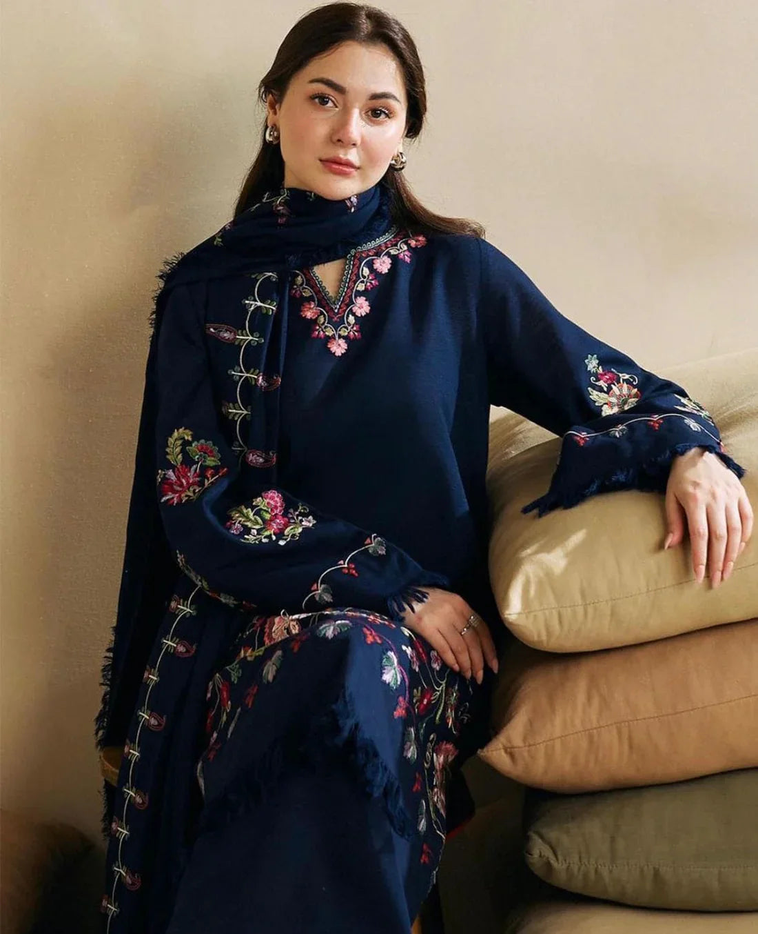 Woman wearing a navy blue embroidered outfit sitting on beige cushions against a beige wall.
