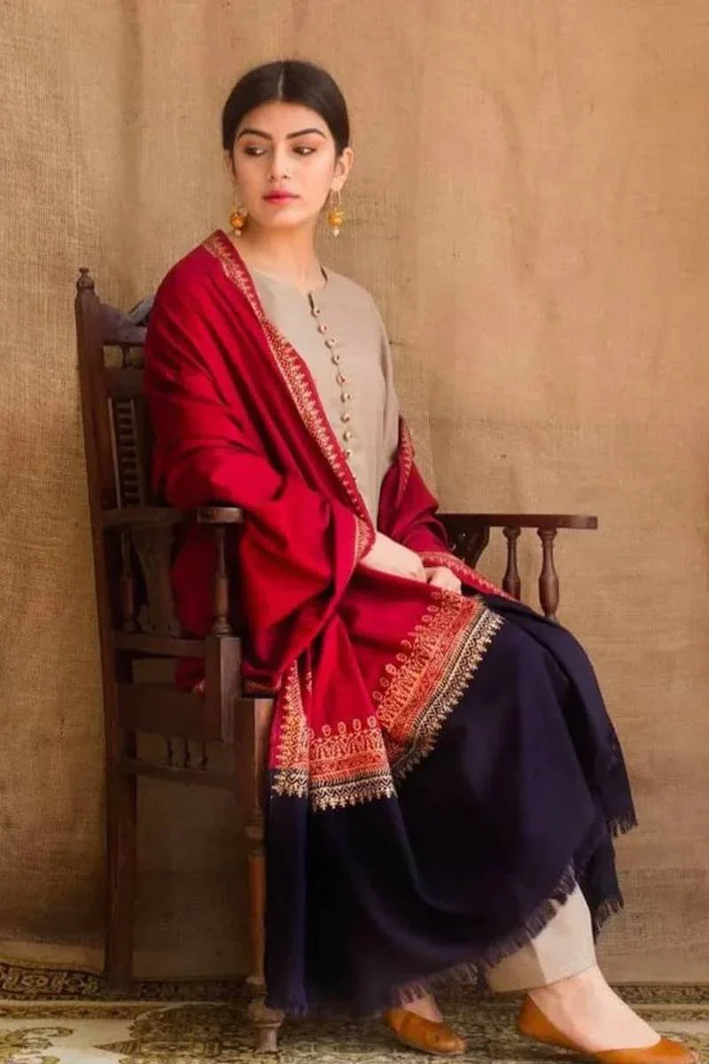 Woman wearing a red and gold embroidered dupatta over a dark blue outfit, sitting on a wooden chair against a beige wall.